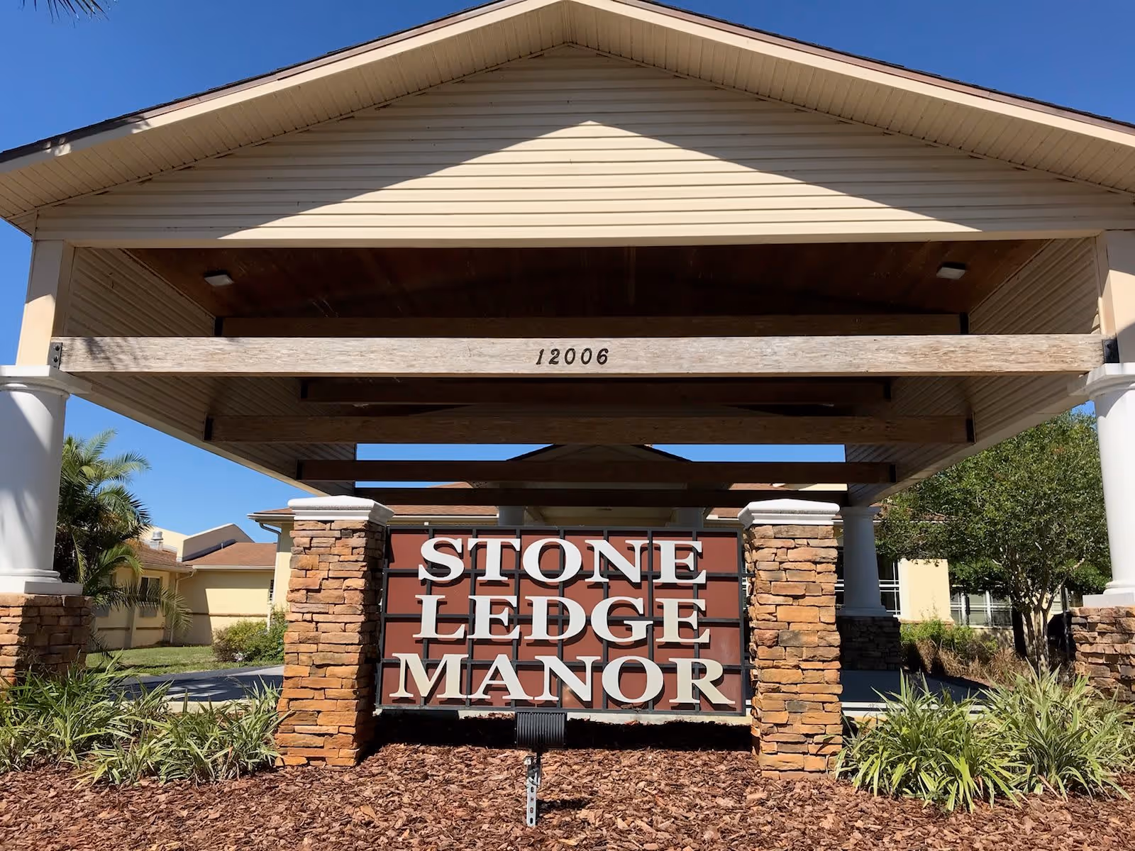Entrance sign for Stone Ledge Manor Senior Living under a covered structure with stone pillars and a clear blue sky in the background.