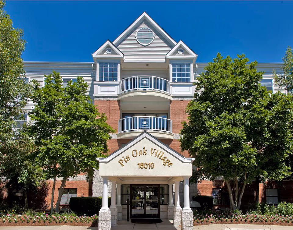 Multi-story senior living building with brick facade, balconies, and a covered entrance labeled Pin Oak Village.
