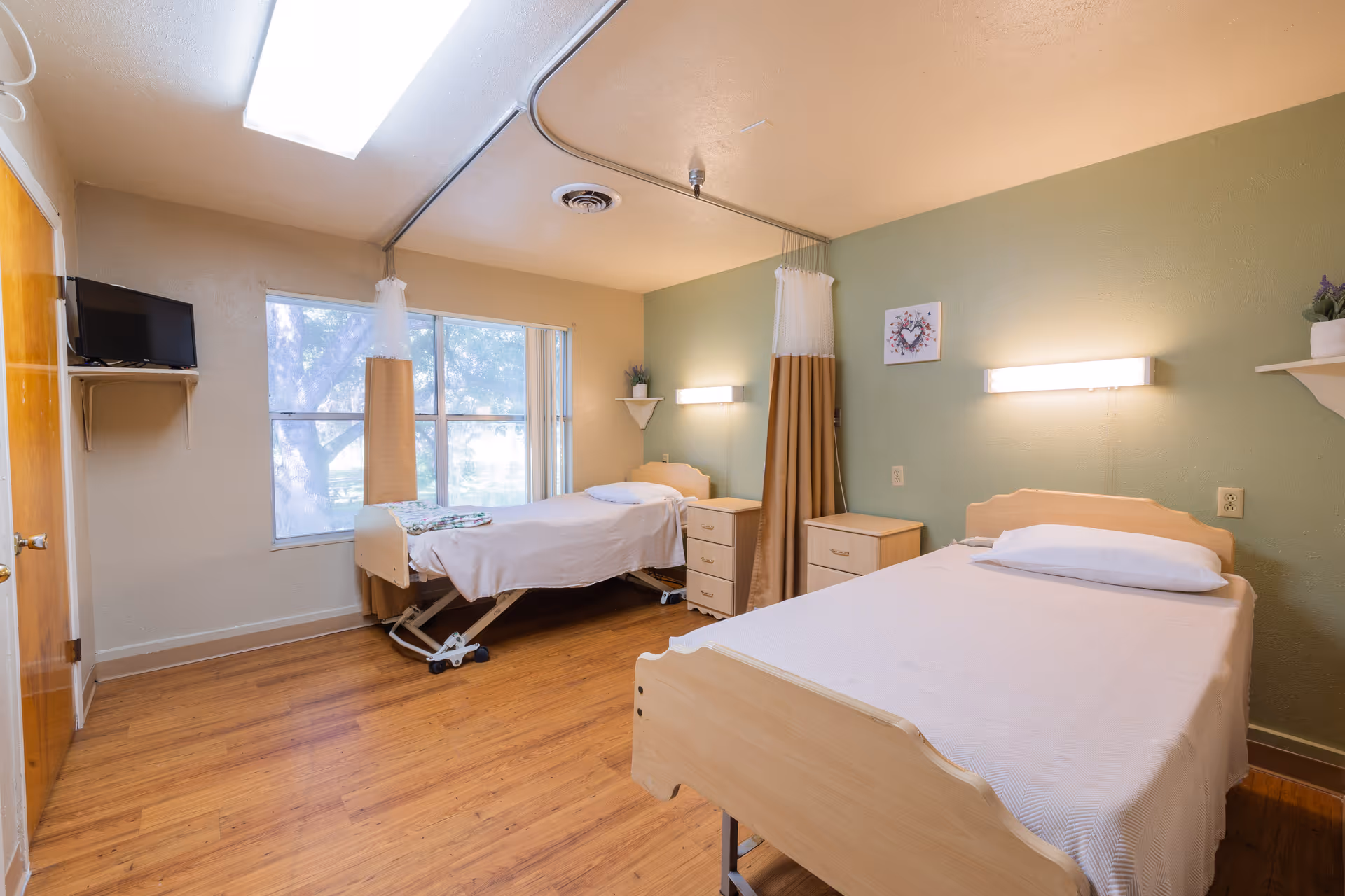 A nursing facility room with two single beds, each with white bedding and a pillow. The room has wooden flooring, a large window letting in natural light, a small TV mounted on the wall, bedside tables, and a curtain divider between the beds. The walls are painted green and beige, with wall-mounted lights above each bed and a small decorative picture on the wall.