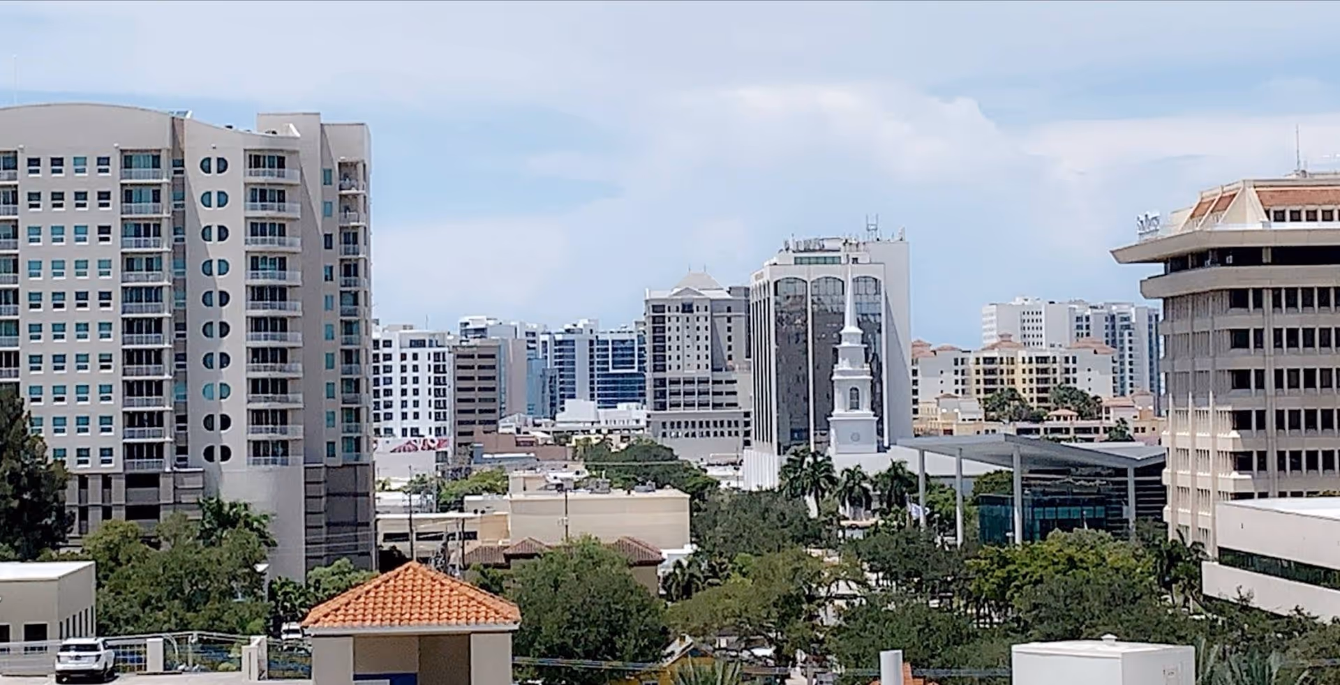 City skyline with tall modern buildings, trees, and a blue sky.