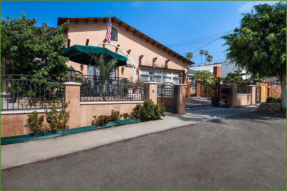 Exterior front of a stucco building with a gated entrance, patio seating under a green umbrella and an American flag.
