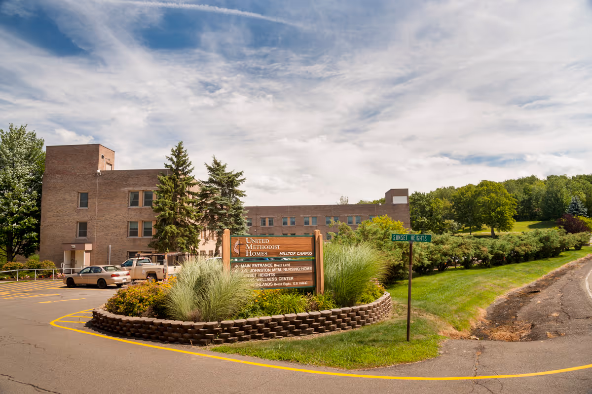 Exterior view of the United Methodist Homes Hilltop Campus building with a parking lot, landscaped greenery, and a sign displaying the facility's name and directions. The sky is partly cloudy and there is a street sign labeled 'Sunset Heights'.