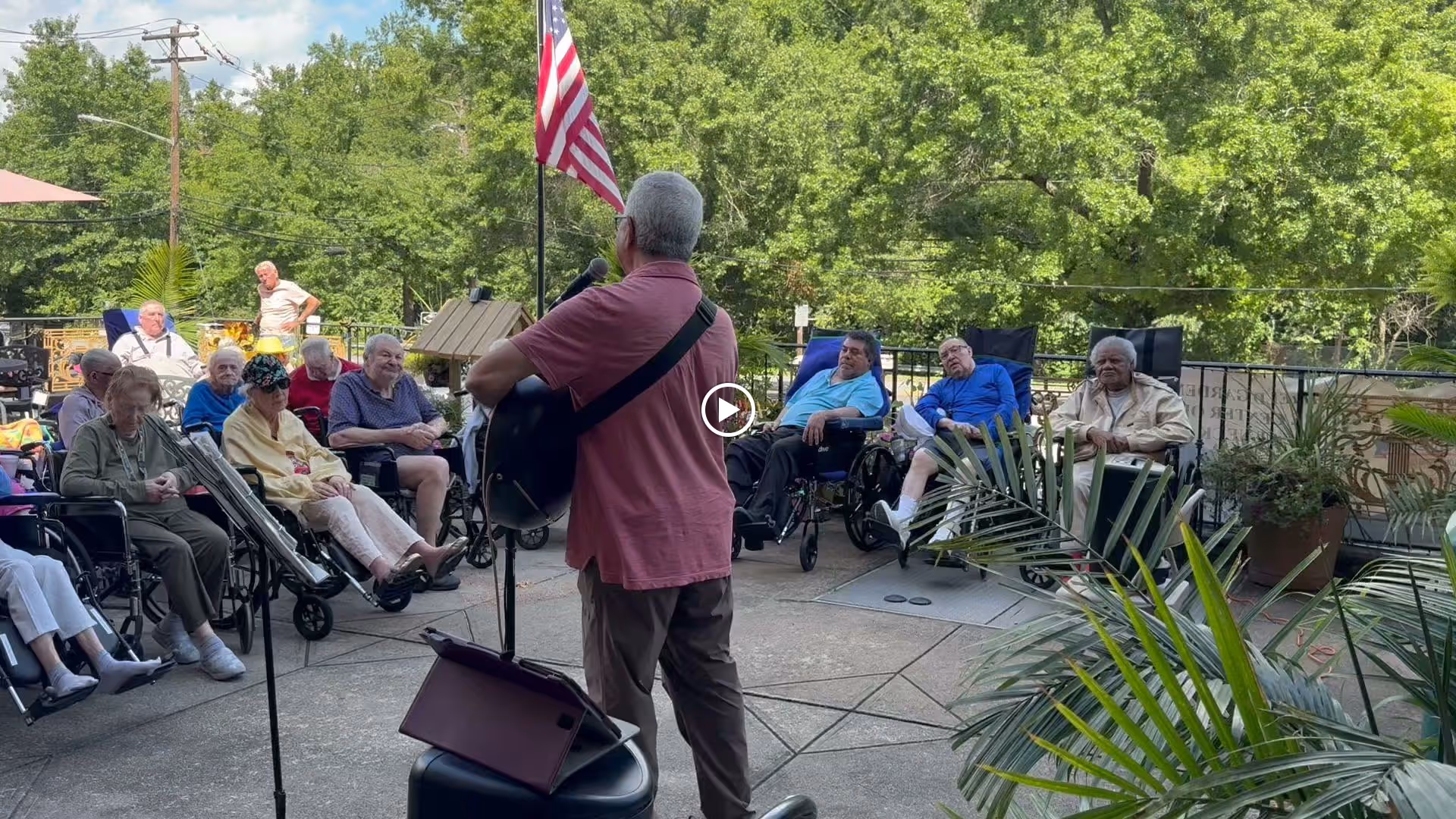 An outdoor patio area at a senior living facility where a man is playing guitar and singing to a seated audience of elderly residents, many in wheelchairs. The setting is surrounded by greenery and an American flag is visible in the background.