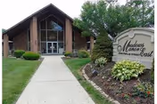 Brick senior living facility entrance with glass doors, curved Meadows Manor East sign, and landscaping.