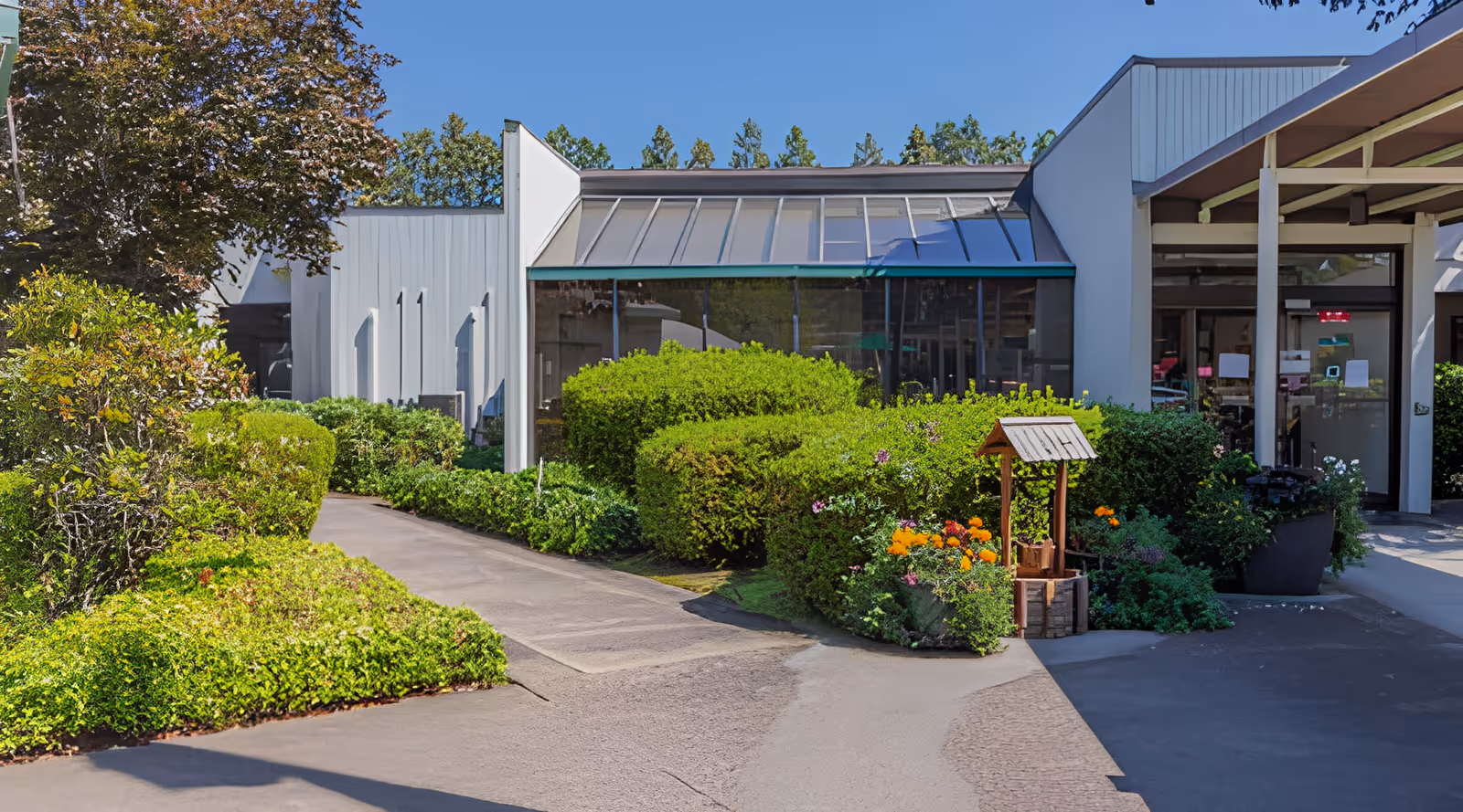 Exterior front entrance of a nursing and rehabilitation center with a landscaped walkway, shrubs, glass windows, and a small decorative wooden well.
