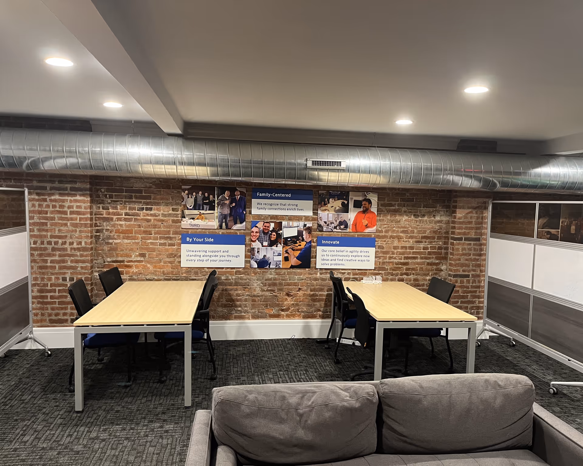 Interior meeting room with two tables, chairs, brick wall, and wall posters.