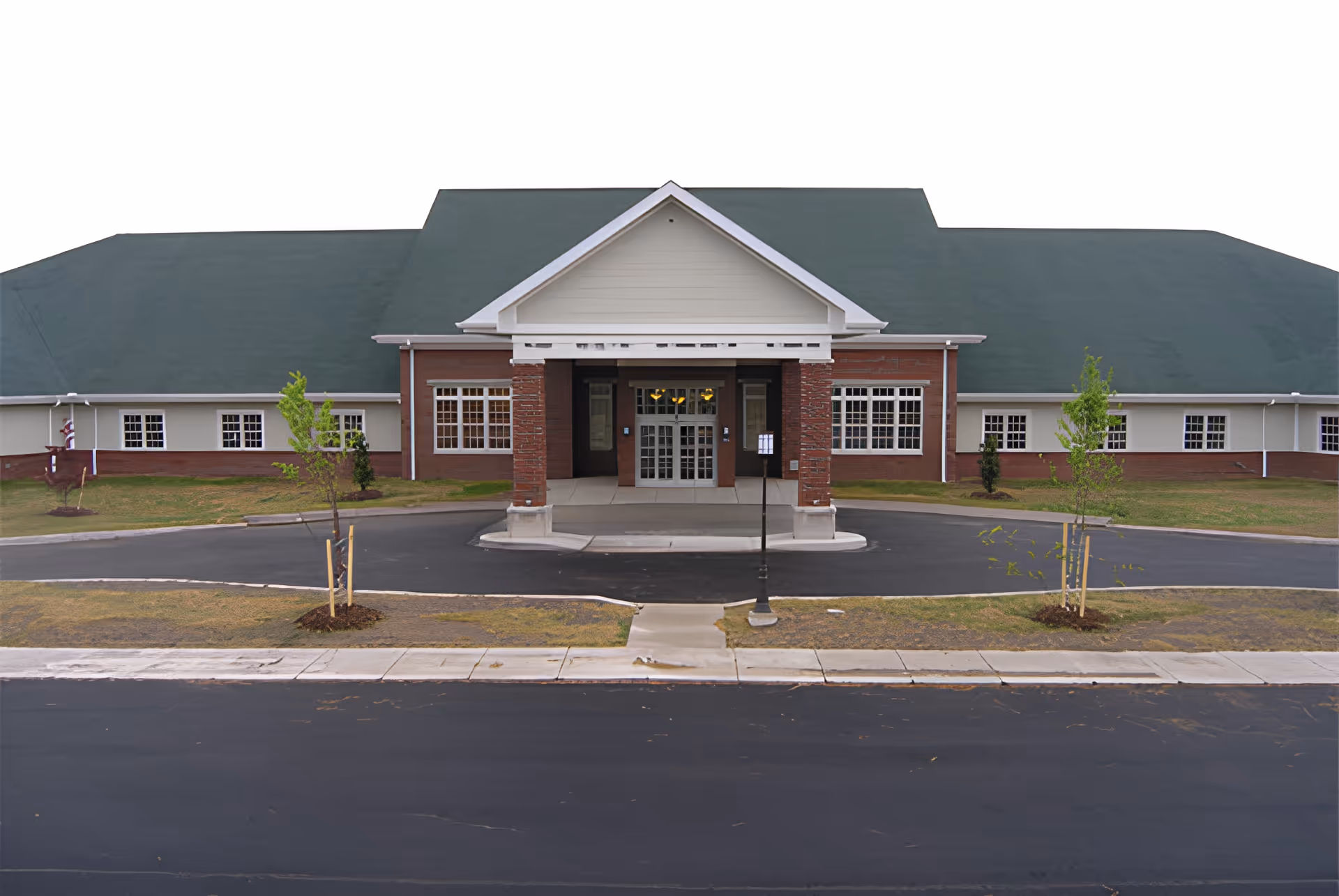 Front exterior view of a single-story building with a green roof and brick and beige siding. The entrance has a covered porch supported by brick columns, with glass double doors. There are small trees and a paved driveway in front of the building.