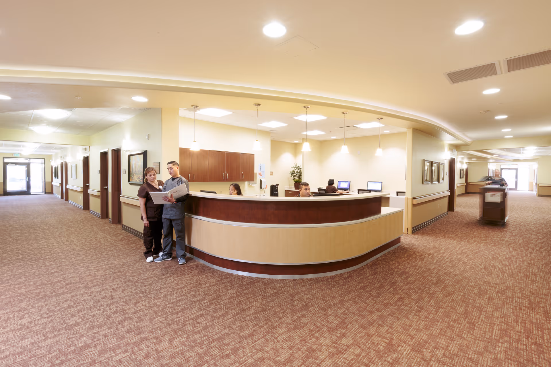A wide, well-lit hallway in a senior living facility with a curved reception desk staffed by two people working on computers. Two healthcare workers stand near the desk reviewing a document. The hallway has carpeted floors, light-colored walls, and multiple doors along the corridor. A housekeeping cart is visible further down the hallway.