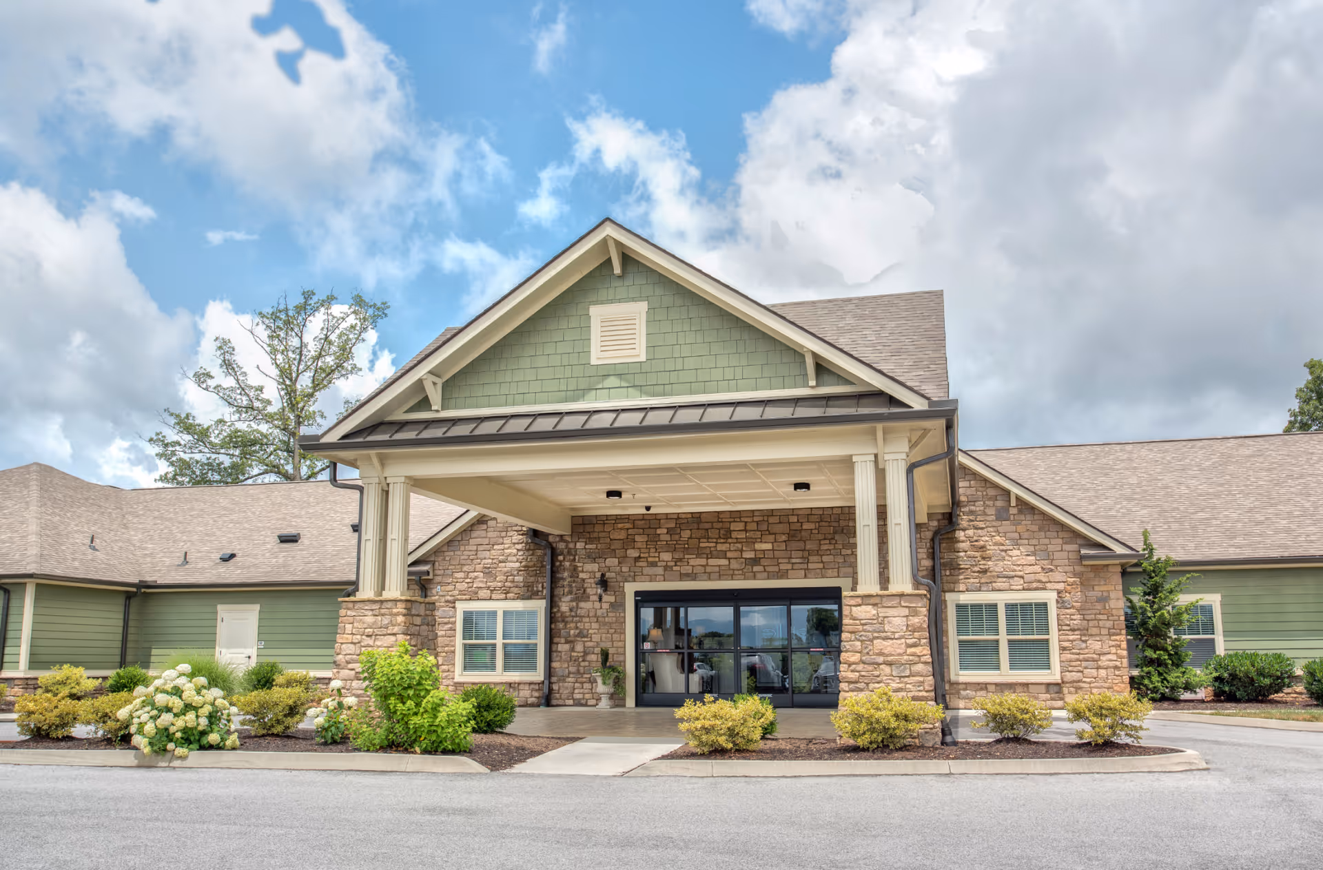 Covered front entrance of a low-rise stone and green-sided senior living building with landscaping under a partly cloudy sky.