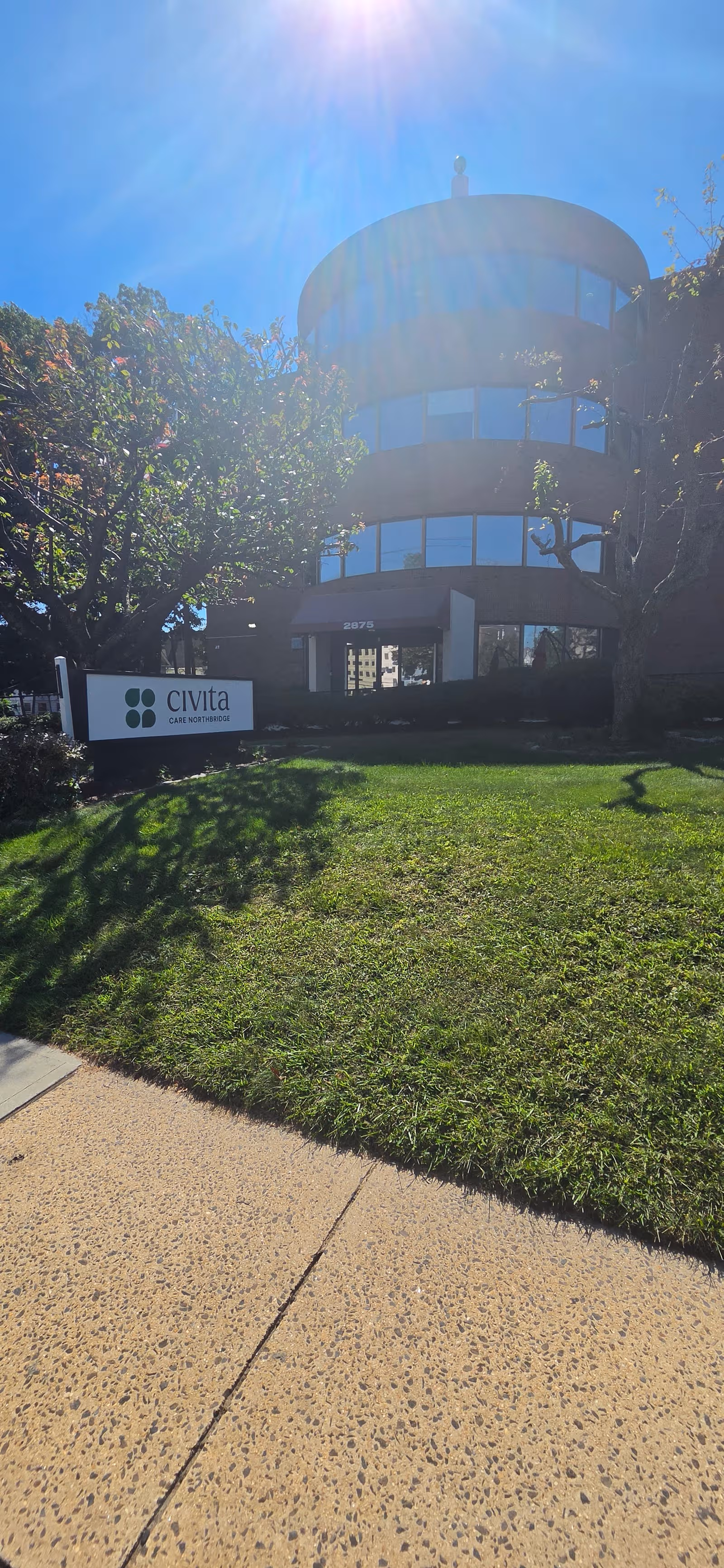 Exterior view of a circular glass-front senior care center with a sign by the lawn