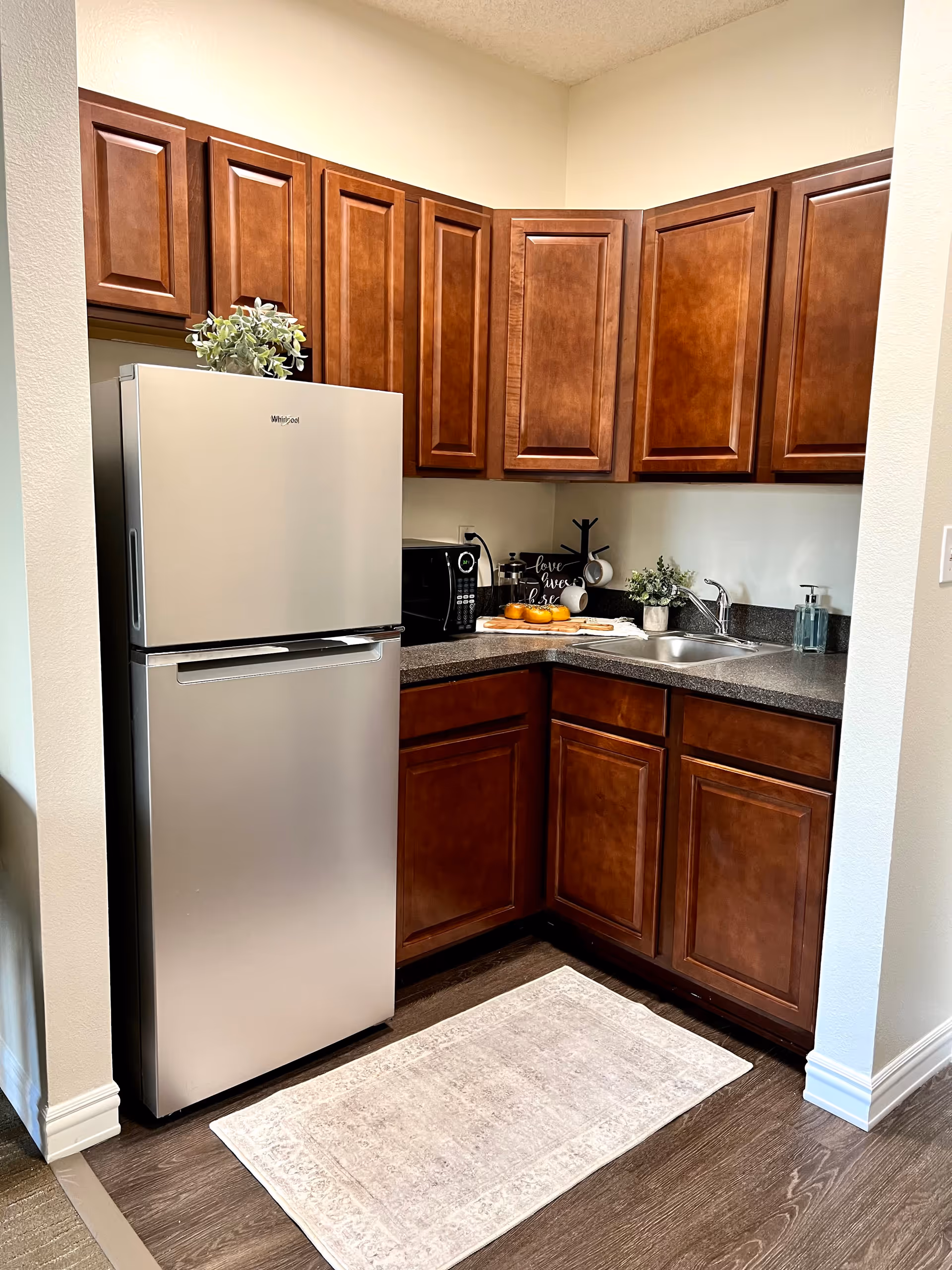A small kitchen area with dark wooden cabinets, a stainless steel Whirlpool refrigerator, a microwave, a sink with a faucet, and a countertop with a cutting board holding oranges and a decorative sign. There is a light-colored rug on the wooden floor in front of the cabinets.