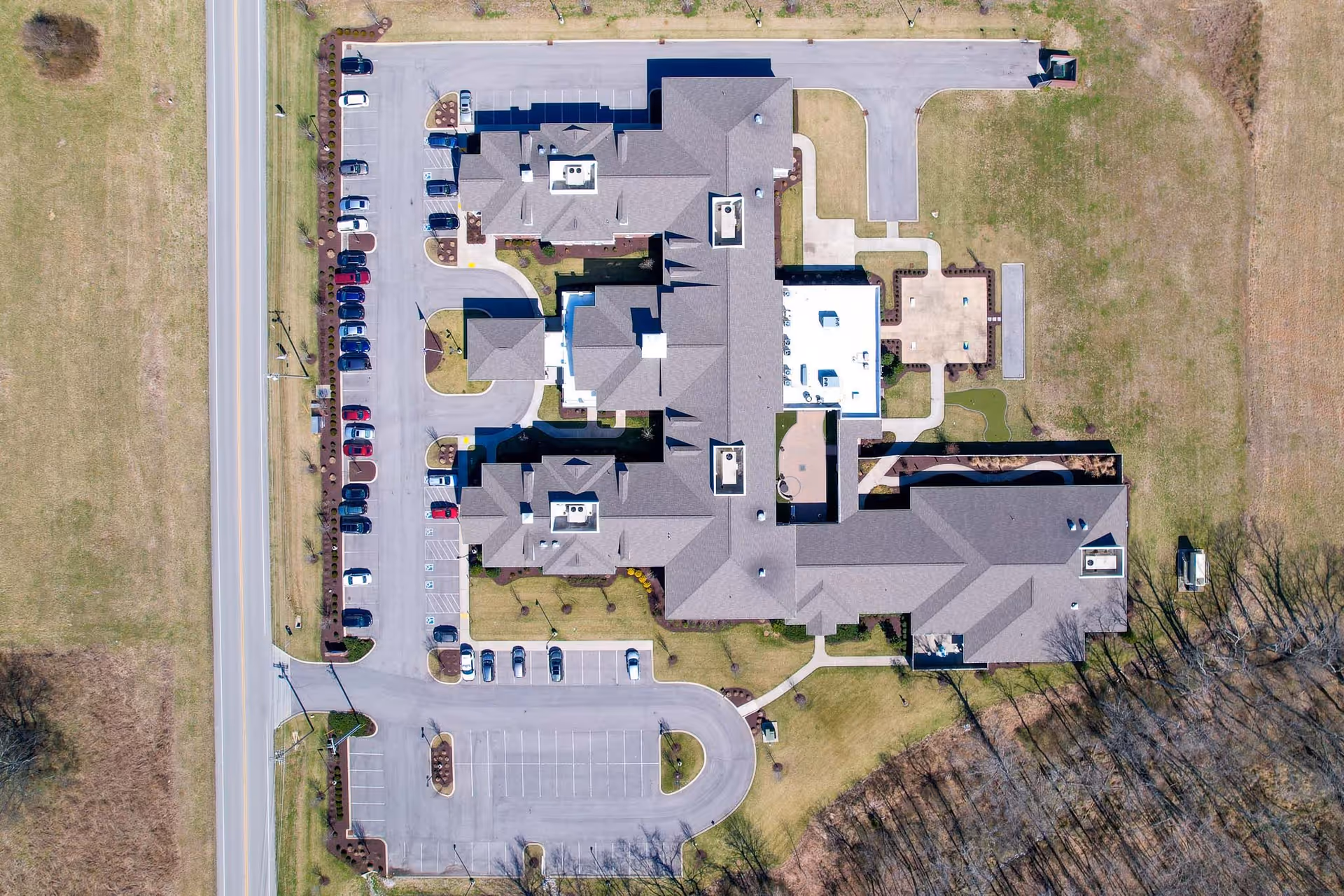 Aerial view of The Goldton at Spring Hill facility showing the building layout, surrounding parking lots with parked cars, adjacent road, and grassy areas around the property.