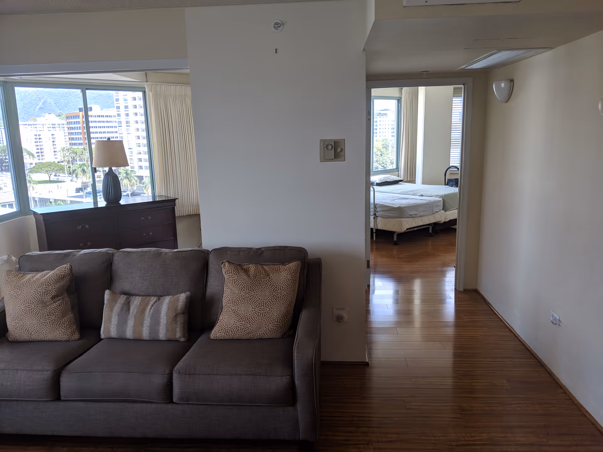 Spacious living area with a gray sofa, dresser, and beds visible through doorway.