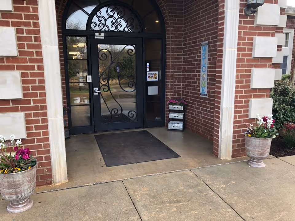 Front entrance of a brick building with a decorative glass door, potted flowers, and a 'Welcome' sign.