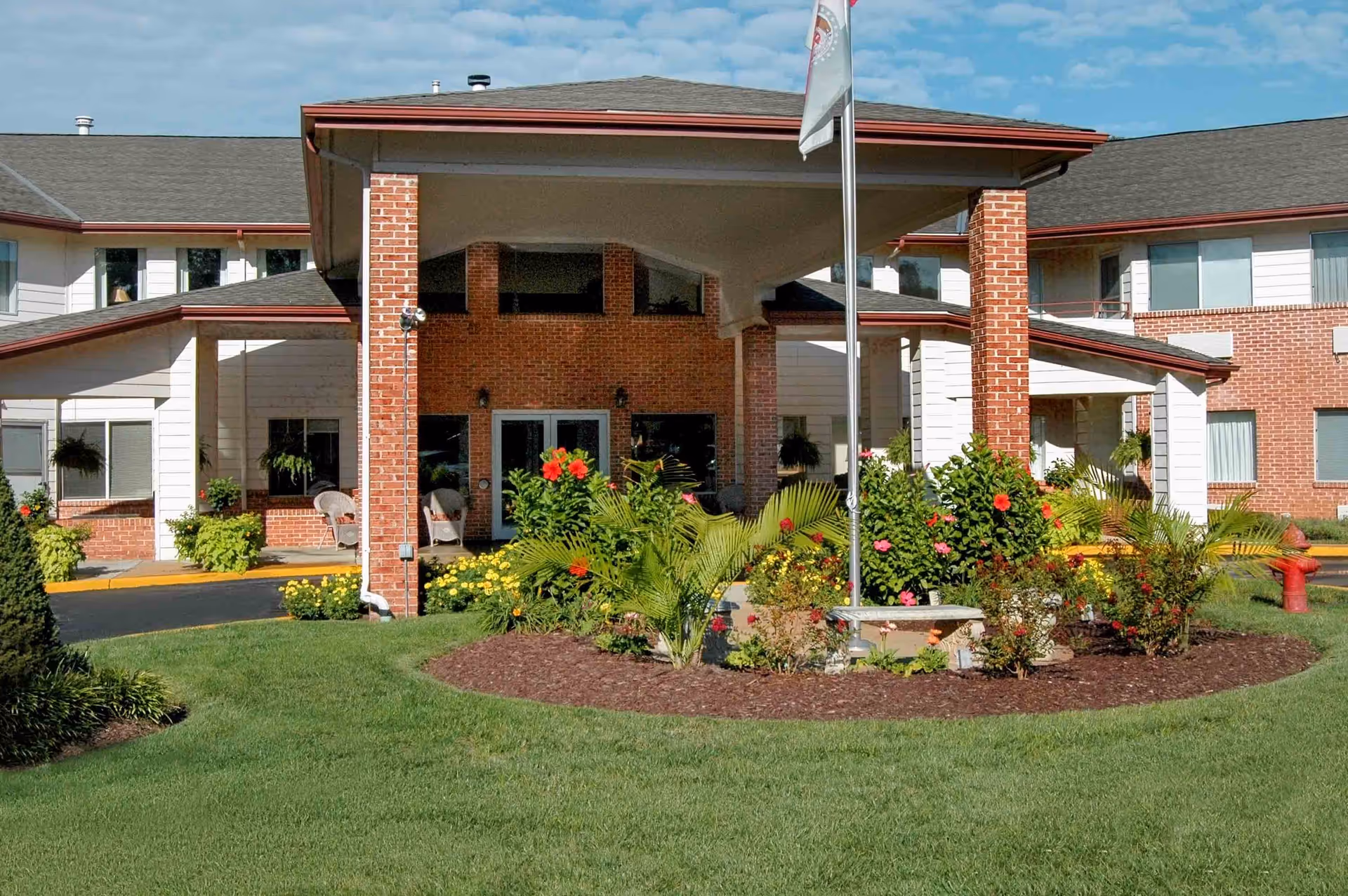Front entrance of Holiday Briarcrest Estates senior living facility with a covered driveway supported by brick pillars, surrounded by well-maintained green lawn and colorful flower beds under a partly cloudy sky.