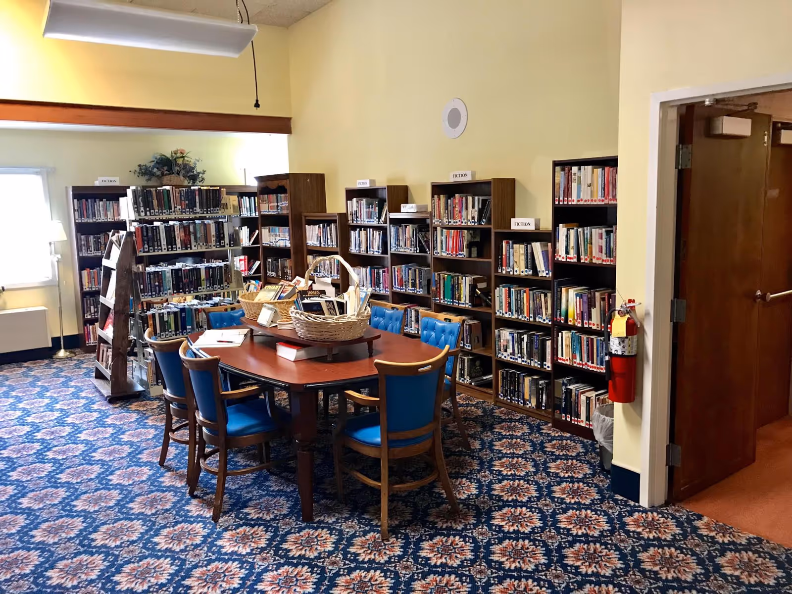 Interior view of a library room with yellow walls and patterned blue carpet. Several wooden bookshelves filled with books labeled 'Fiction' line the walls. A wooden table with blue cushioned chairs surrounds it, and baskets with books are placed on the table. A fire extinguisher is mounted on the wall near an open door.