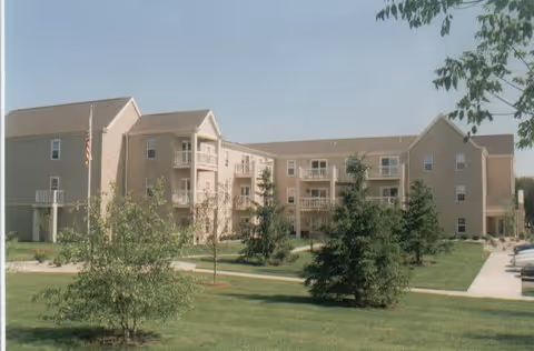 Exterior view of a multi-story senior apartment building with beige siding, balconies, and an American flag on a flagpole. The building is surrounded by green grass, trees, and paved walkways under a clear blue sky.