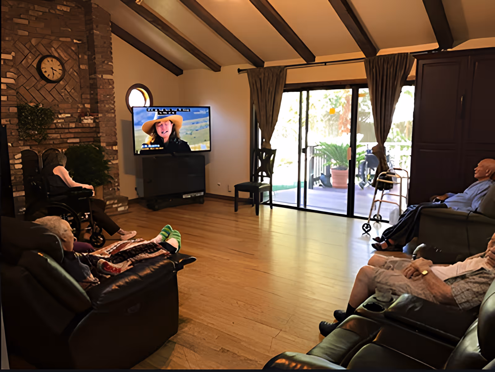 Several elderly residents seated in a communal living room watching television near a brick fireplace and sliding glass doors.