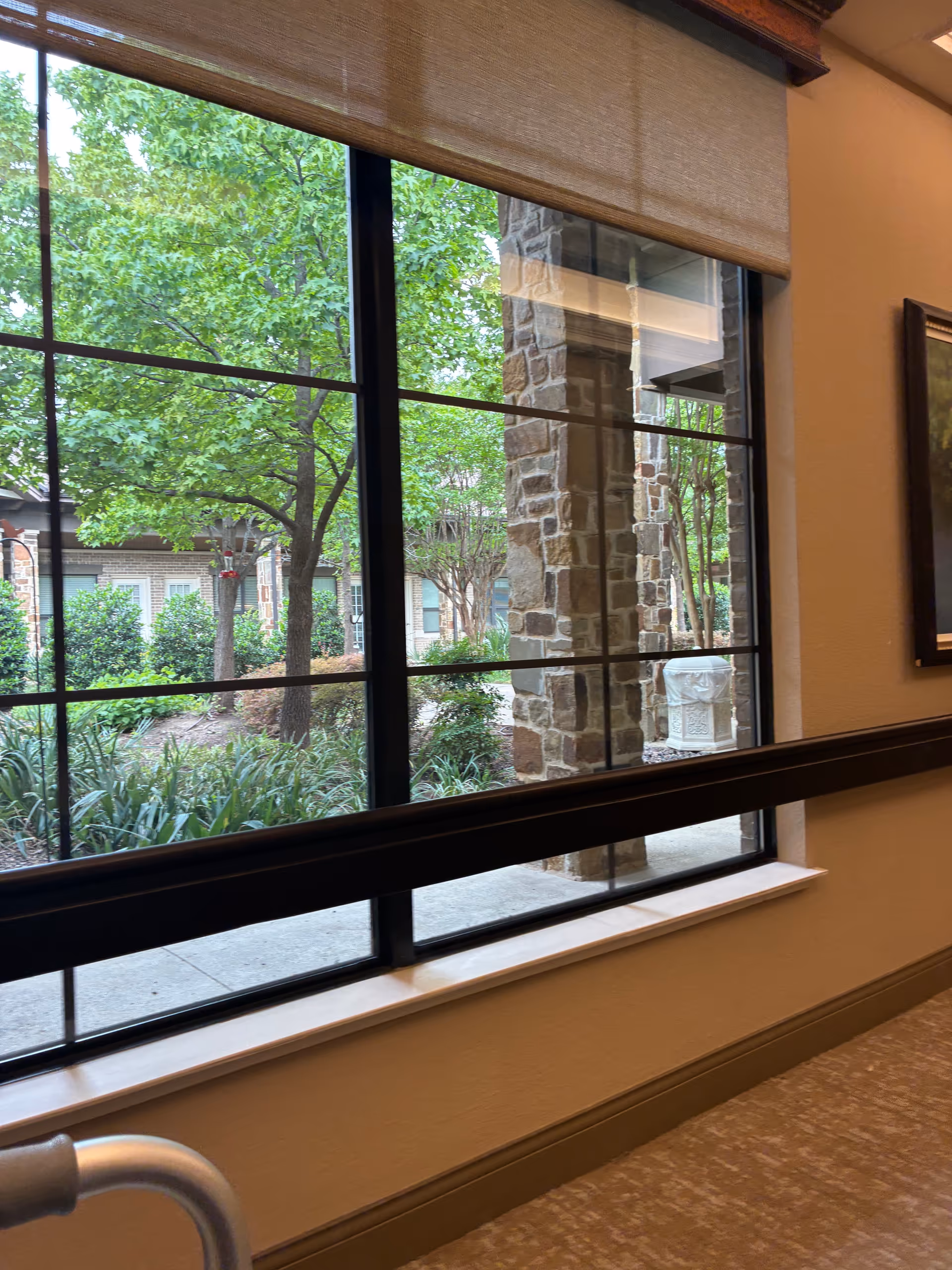 Inside a senior living facility, hallway view through large windows reveals trees and a stone column.