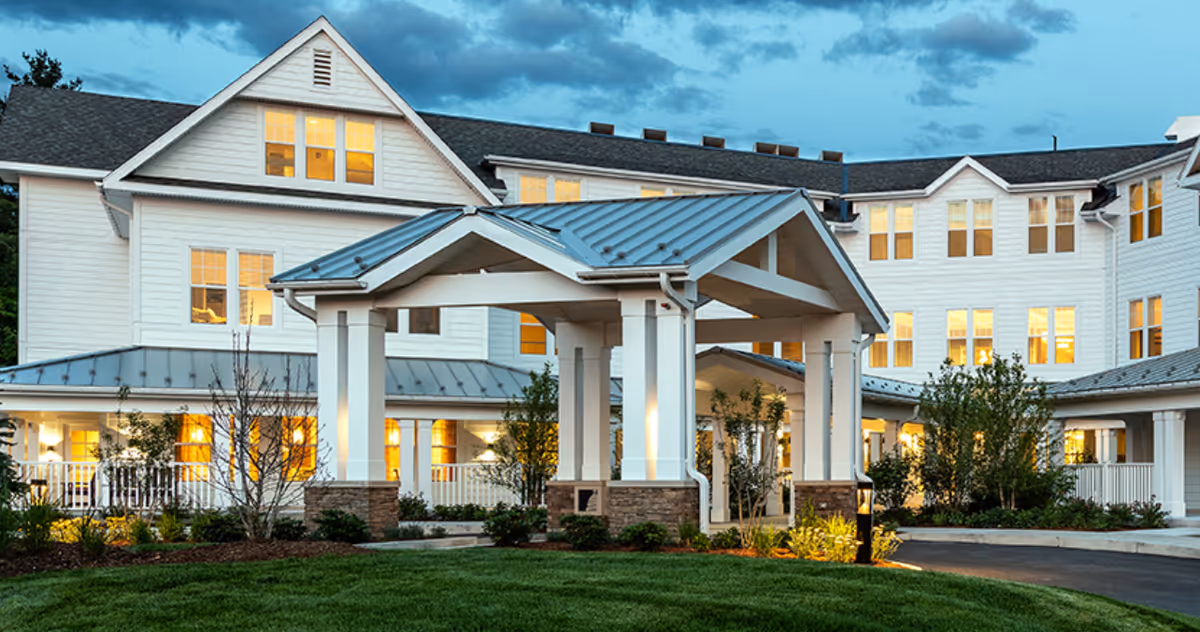 White senior living facility with a covered entrance, illuminated windows, and manicured landscaping at dusk.