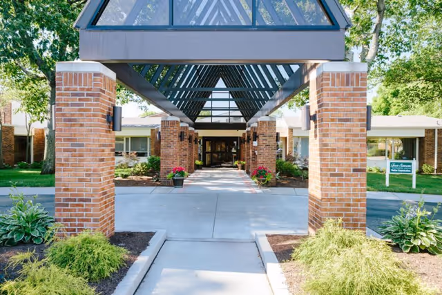Entrance to a residential building with a covered walkway supported by brick pillars and a glass roof. There are plants and flowers along the walkway, and the building is surrounded by trees and greenery.