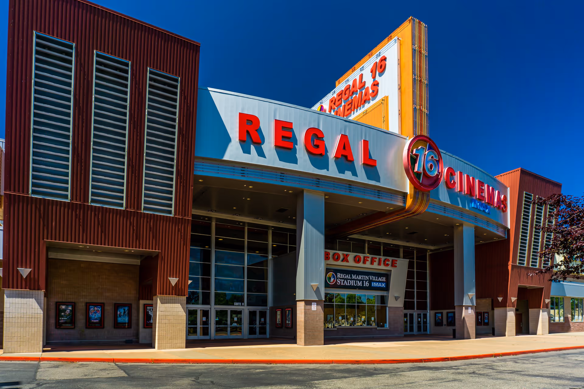 Exterior view of a Regal Cinemas movie theater with a large sign displaying 'REGAL 16 CINEMAS' and a box office window below. The building has a modern design with red and beige accents under a clear blue sky.