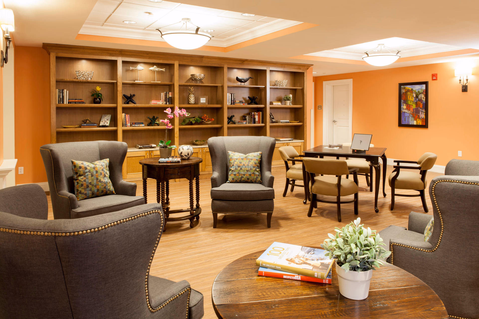 A cozy senior living facility common area with orange walls, wooden flooring, and a large wooden bookshelf filled with books and decorative items. The room features several gray upholstered armchairs with patterned cushions, a round wooden table with books and a potted plant, and a rectangular table with four beige chairs. The ceiling has recessed lighting and two ceiling light fixtures, and a colorful framed painting hangs on the wall.