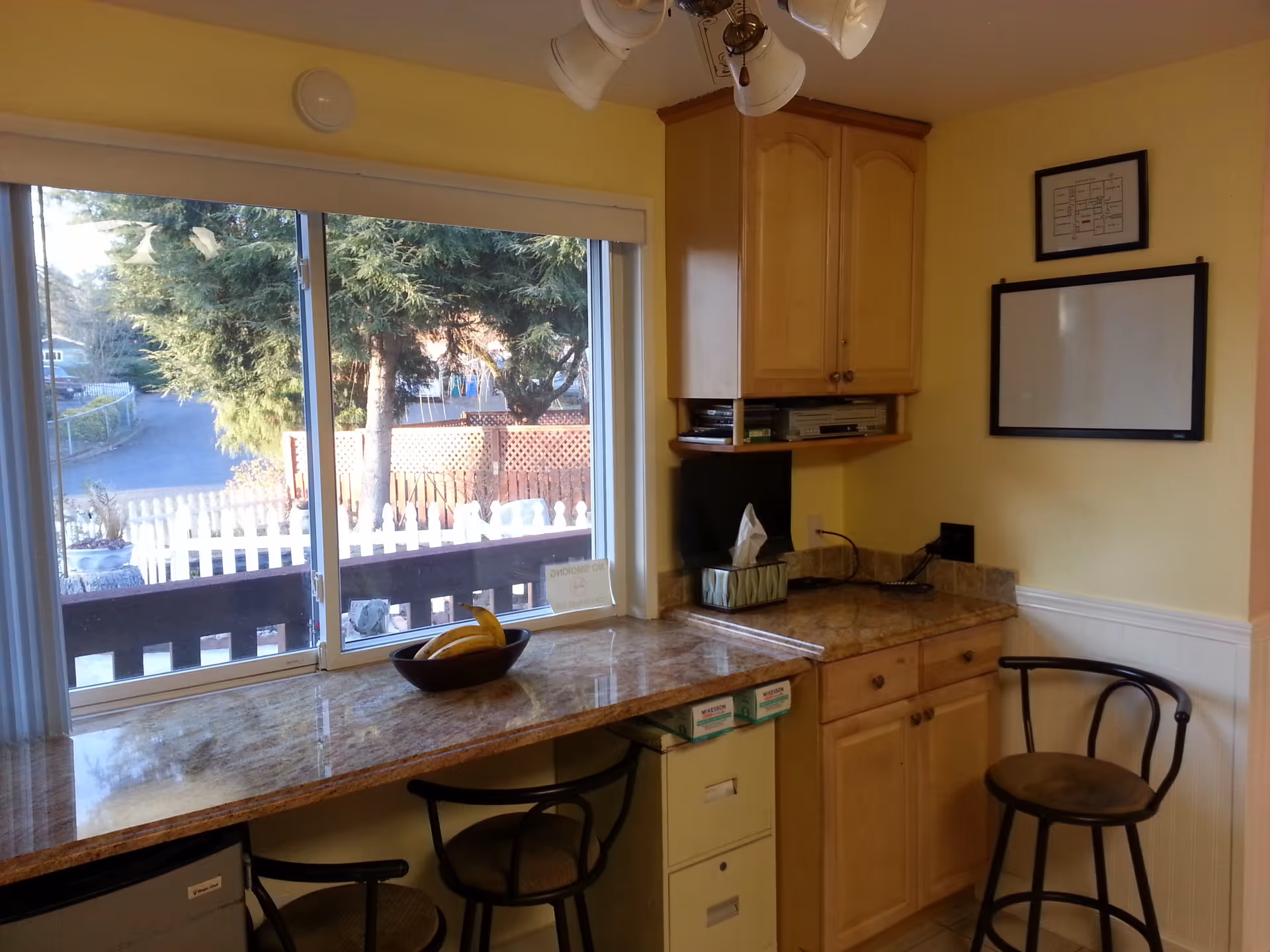 Sunlit kitchen counter with bar stools, wooden cabinets, and a large window overlooking a picket fence and trees.