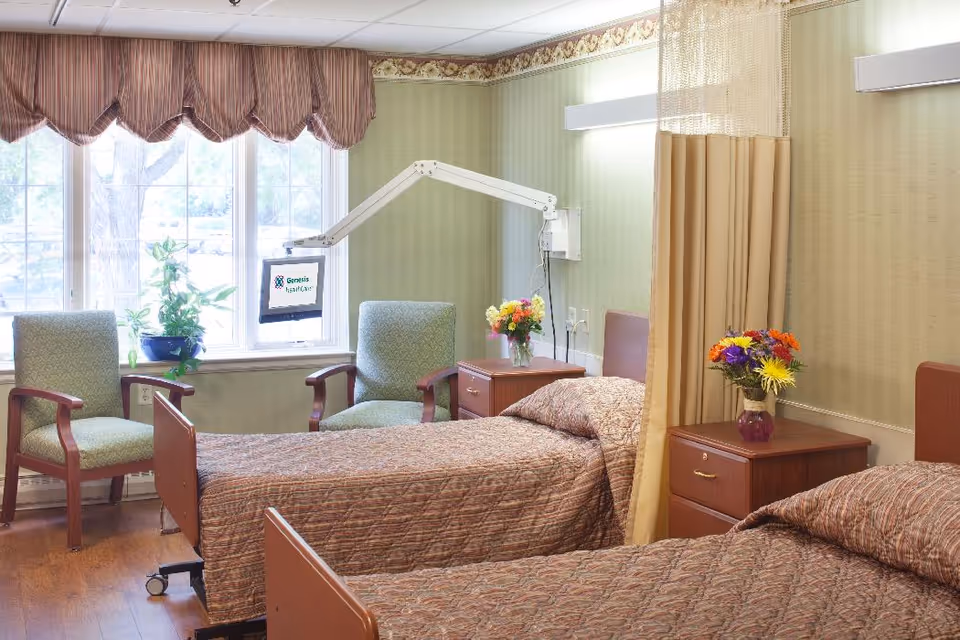 A shared bedroom in a senior living facility with two beds, chairs, and a decorative plant.