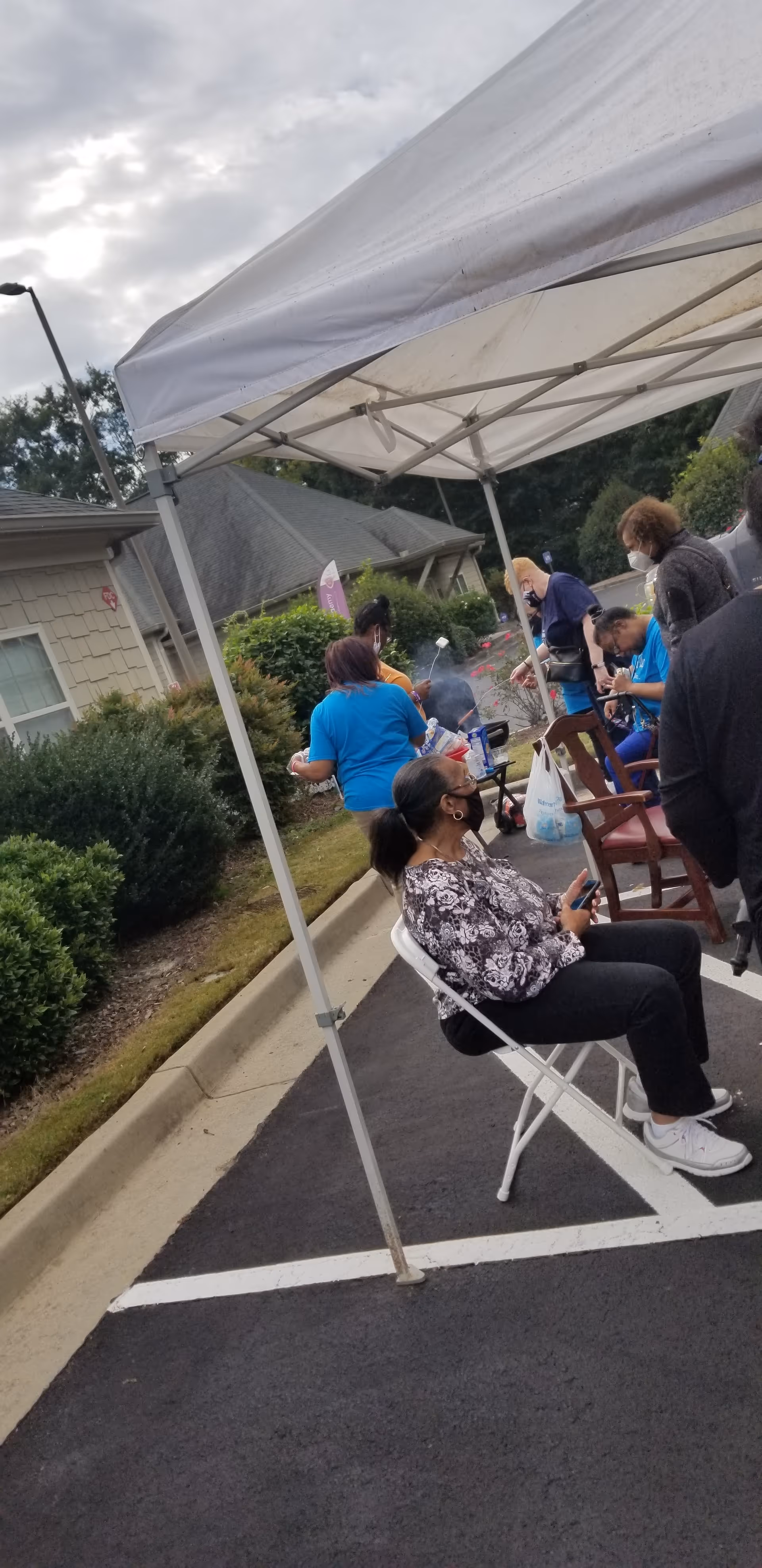 Outdoor gathering under a white canopy with residents seated and chatting near a snack table.
