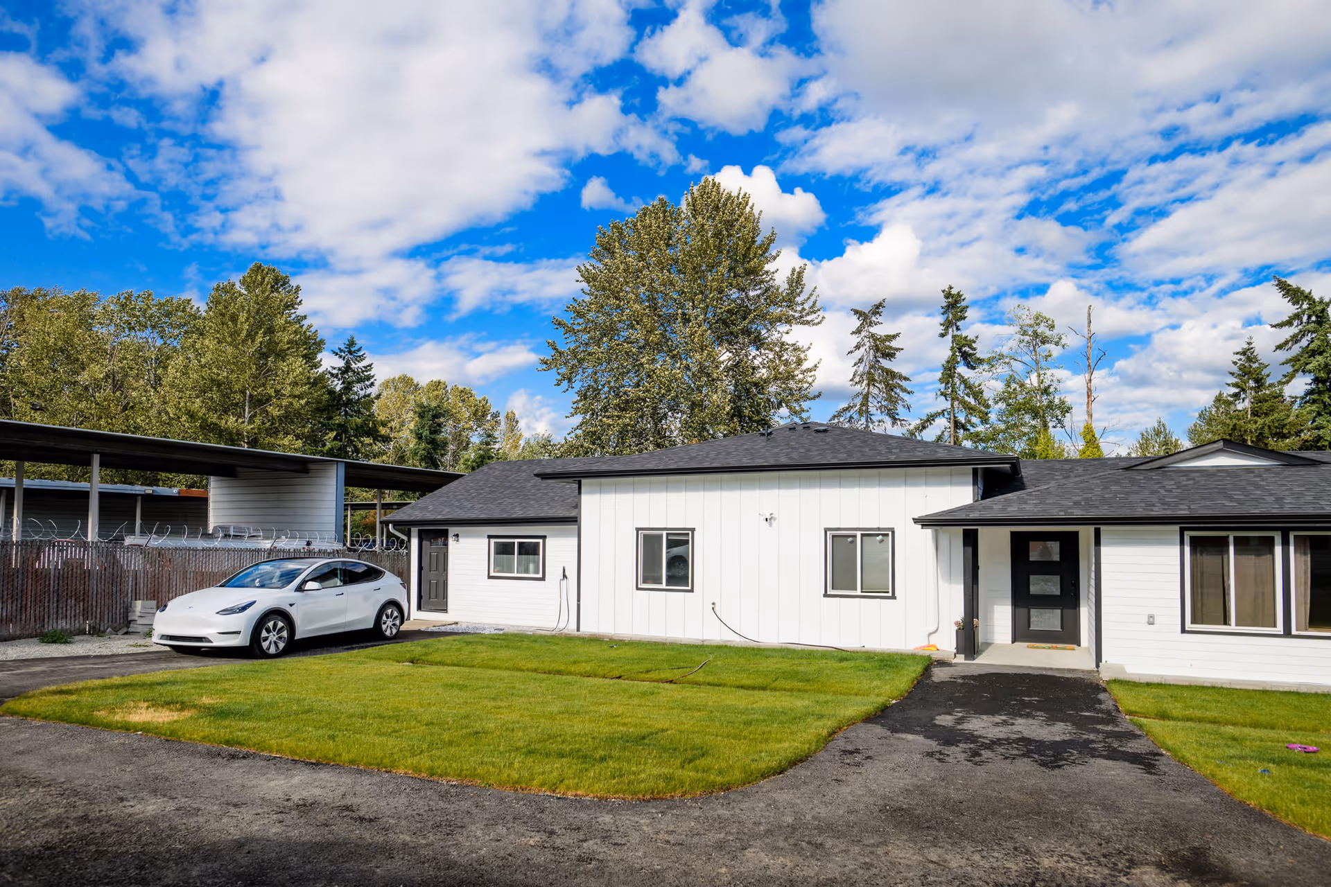 Single-story white facility with black roof, car on driveway, green lawn, and trees behind.