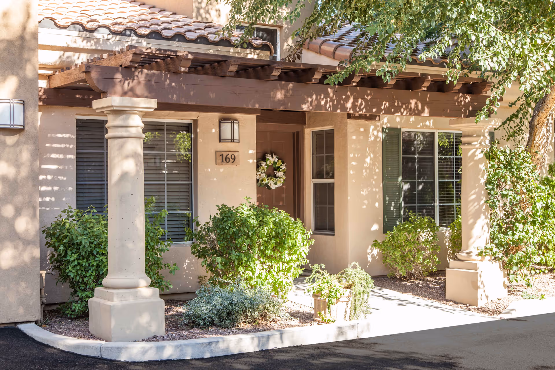 Exterior view of a residential unit entrance at Tuscany at McCormick Ranch featuring a covered porch with wooden beams and columns, a door decorated with a floral wreath, windows with shutters, and surrounding greenery including bushes and small plants.