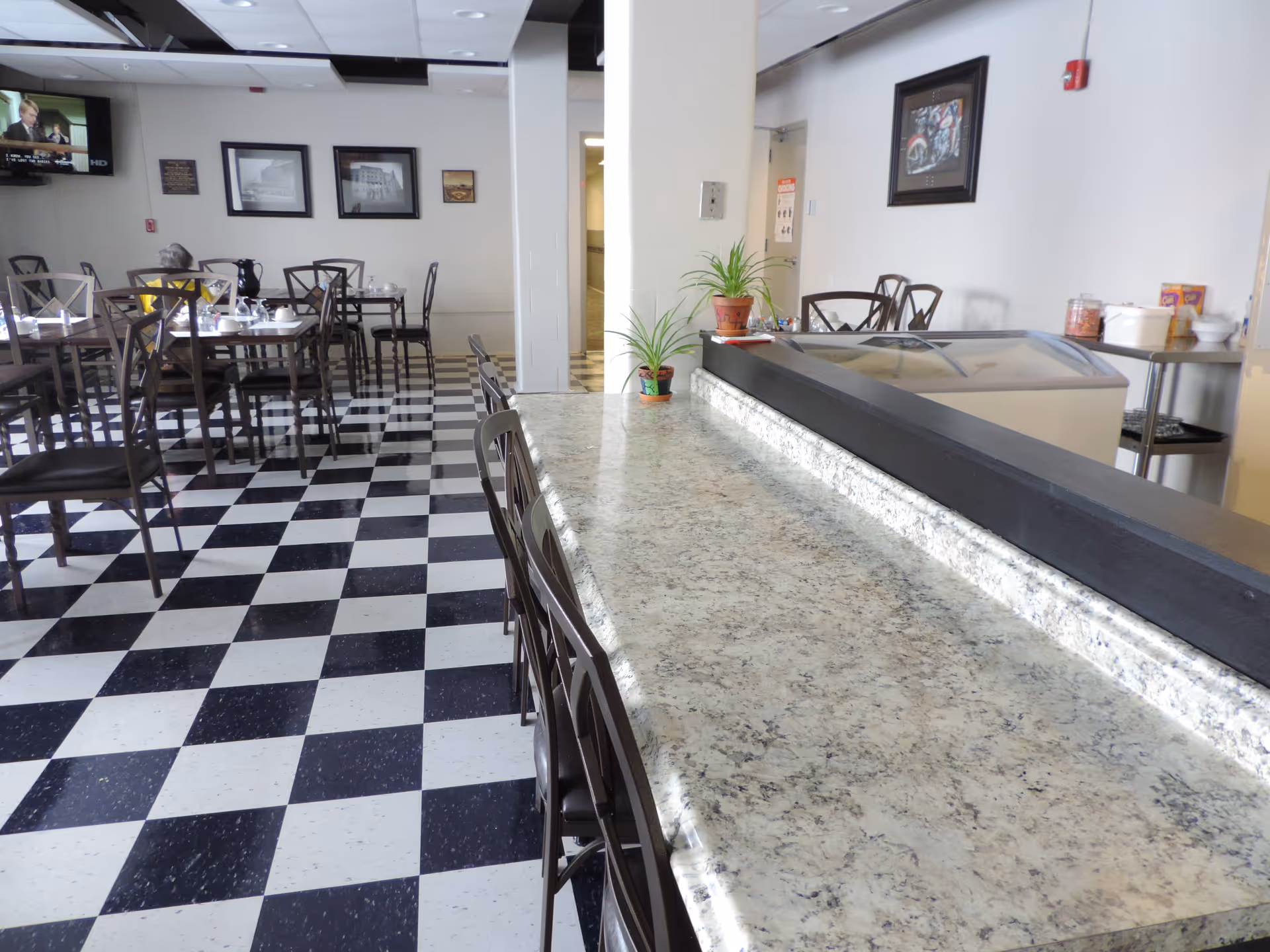 Interior view of a dining area with black and white checkered floor tiles, several tables and chairs arranged for seating, a granite countertop with bar stools in the foreground, potted plants on the counter, framed pictures on the walls, and a television mounted on the wall in the background.