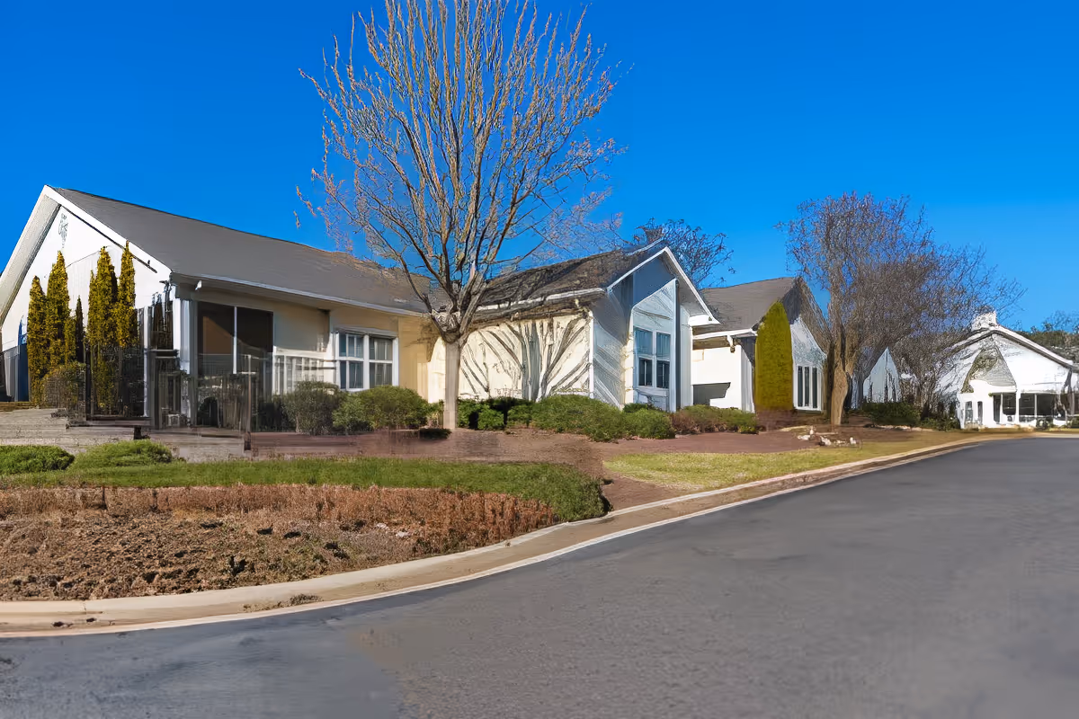 Exterior view of a single-story building with beige walls and a gray roof, surrounded by leafless trees and shrubs under a clear blue sky. The building is adjacent to a paved road with a curb.