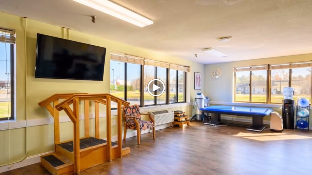 A bright rehabilitation room with large windows letting in natural light. The room features a mounted flat-screen TV, a wooden therapy step platform, a floral upholstered chair, a blue therapy table, a water cooler, and exercise equipment. The walls are painted light yellow and light blue, and there is a clock on the wall.