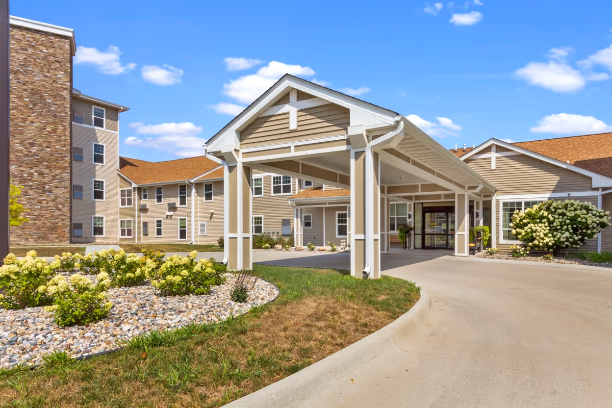 Exterior view of Mill Pond senior living facility showing the main entrance with a covered driveway, beige siding, stone accents, and landscaped garden beds with flowering bushes under a blue sky with scattered clouds.
