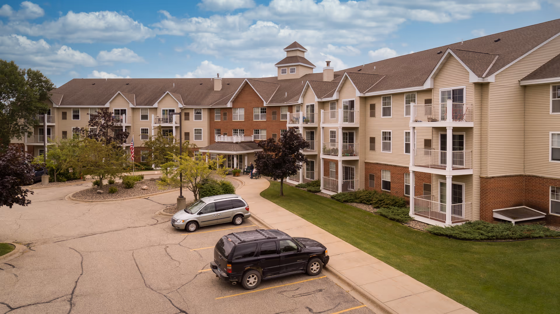 Three-story beige senior living building with balconies, a circular driveway, and parked cars.