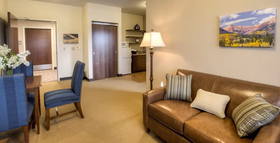 Interior view of a senior living facility room featuring a brown leather sofa with three pillows, a standing lamp, a dining table with blue upholstered chairs, and a small kitchenette area with a refrigerator and microwave. A landscape painting hangs on the wall above the sofa.