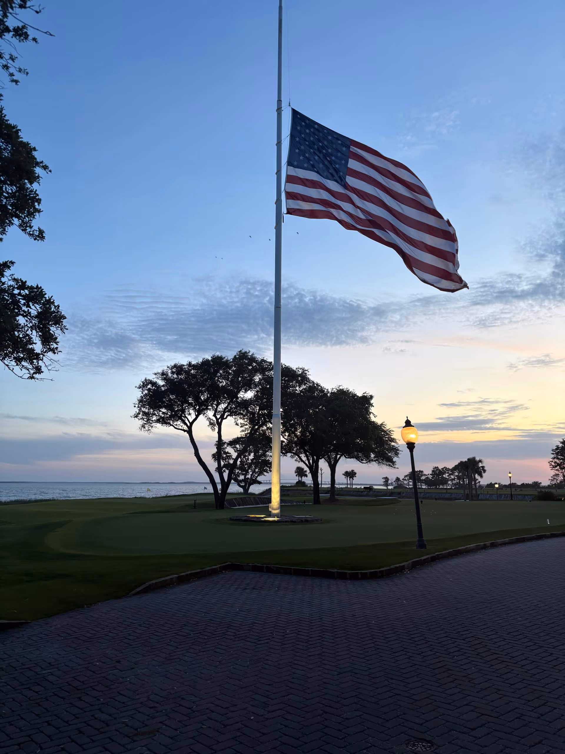 A flagpole with a waving American flag stands in a waterfront outdoor park at dusk.