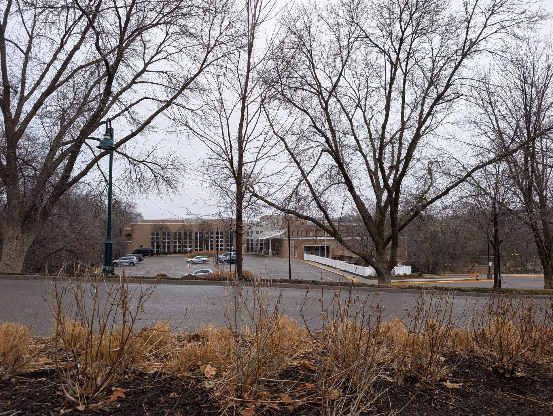 Brick rehabilitation institute building and entrance seen across a parking lot framed by leafless trees and roadside plantings.