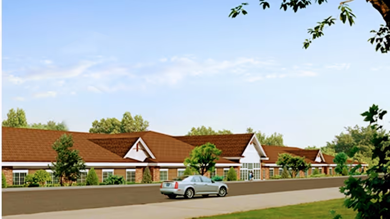 Exterior view of a single-story senior living facility building with a brown roof, white walls, and large windows. There is a silver car parked on the driveway in front of the building, surrounded by green trees and shrubs under a partly cloudy sky.