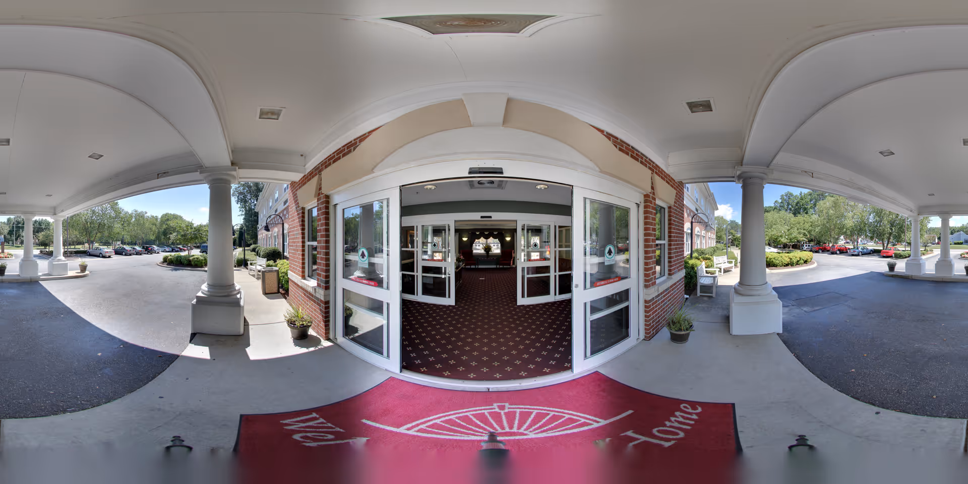 Covered entrance area of a senior living facility with white columns and a red welcome mat in front of automatic sliding glass doors leading inside. The exterior walls are brick, and there are potted plants near the entrance. A parking lot with cars and greenery is visible on both sides under a clear blue sky.