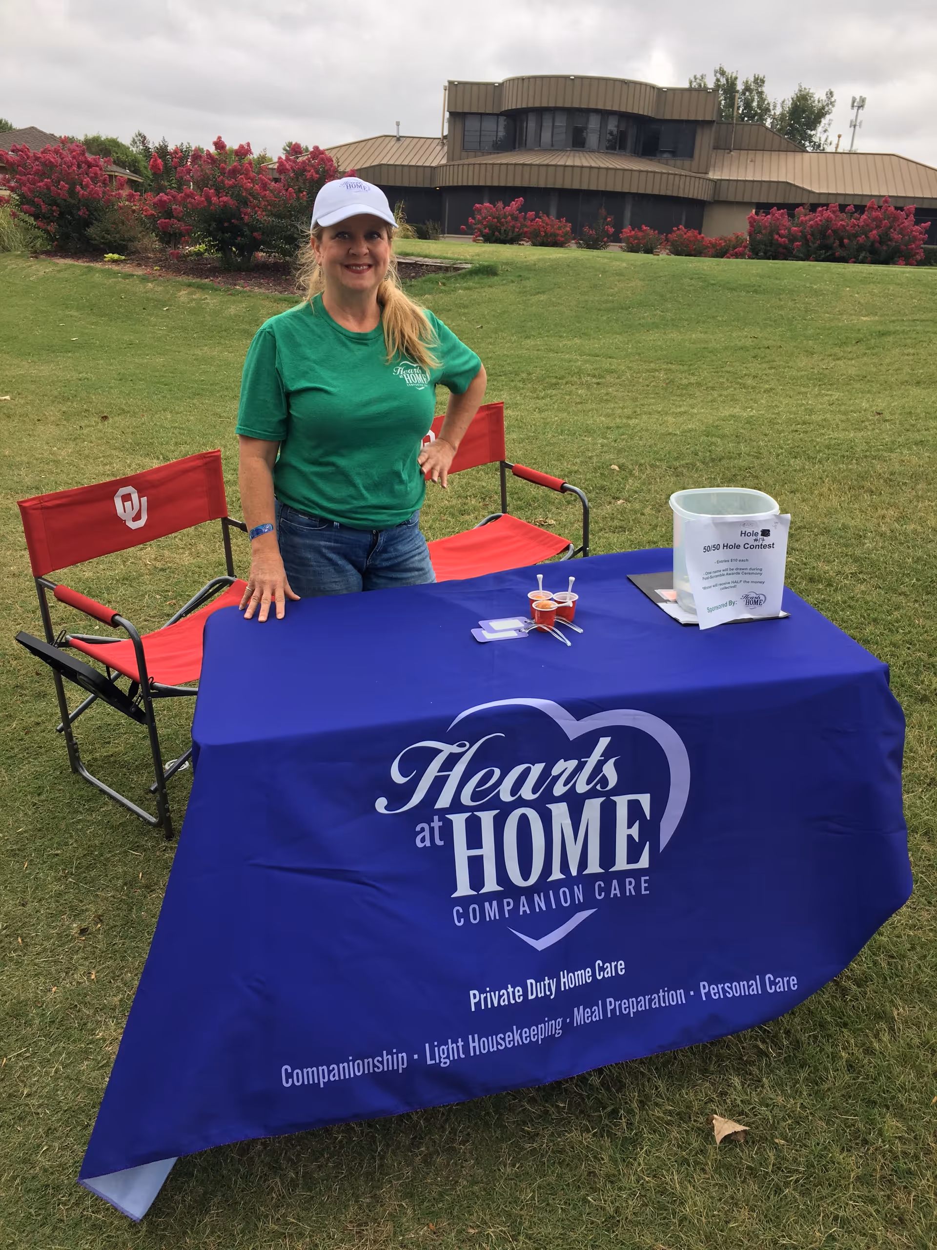 Smiling woman stands beside a Hearts at Home table at an outdoor event.