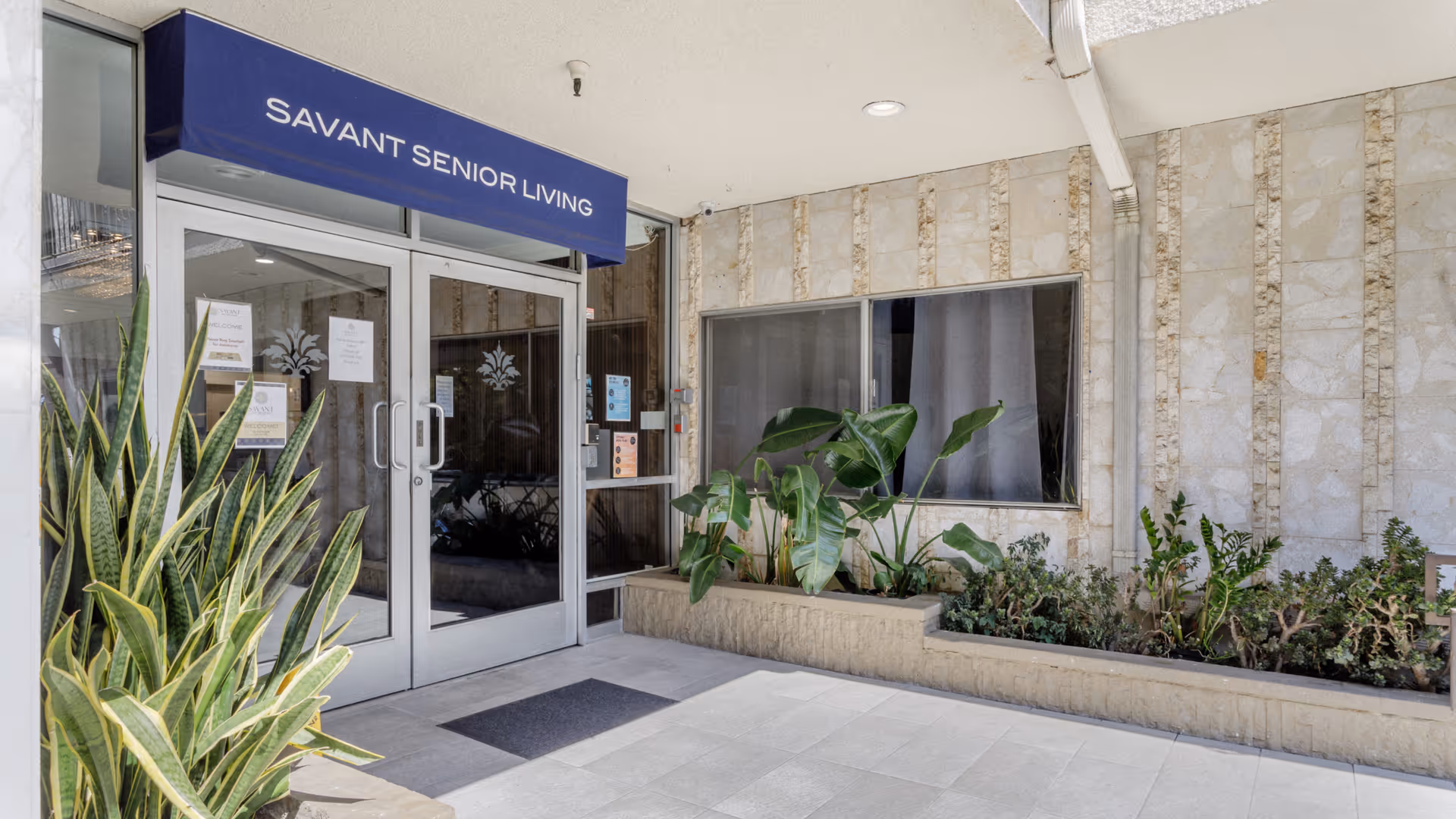 Savant Senior Living entrance with glass doors, blue awning, and potted plants.