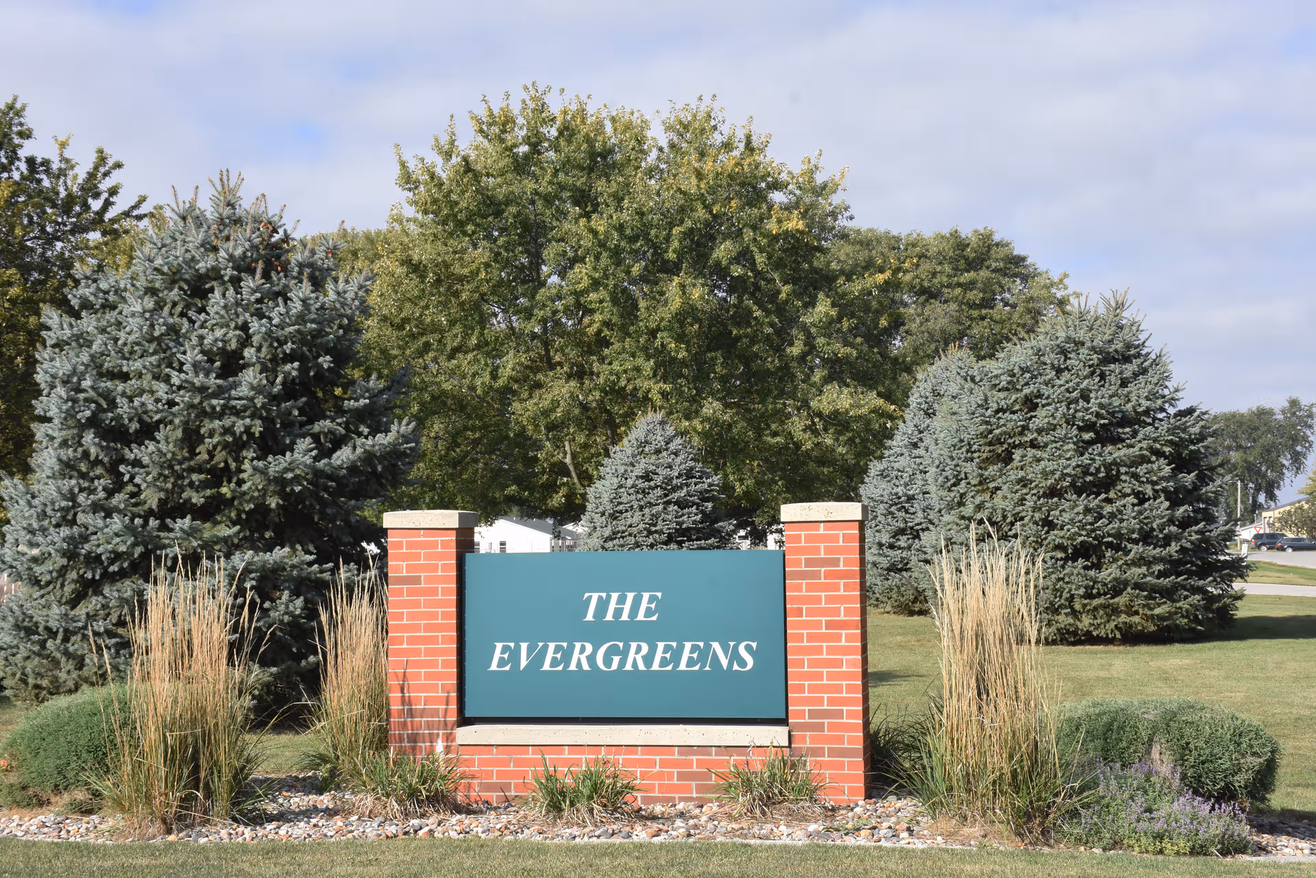 A landscaped entrance sign reads THE EVERGREENS between brick columns and evergreen trees.
