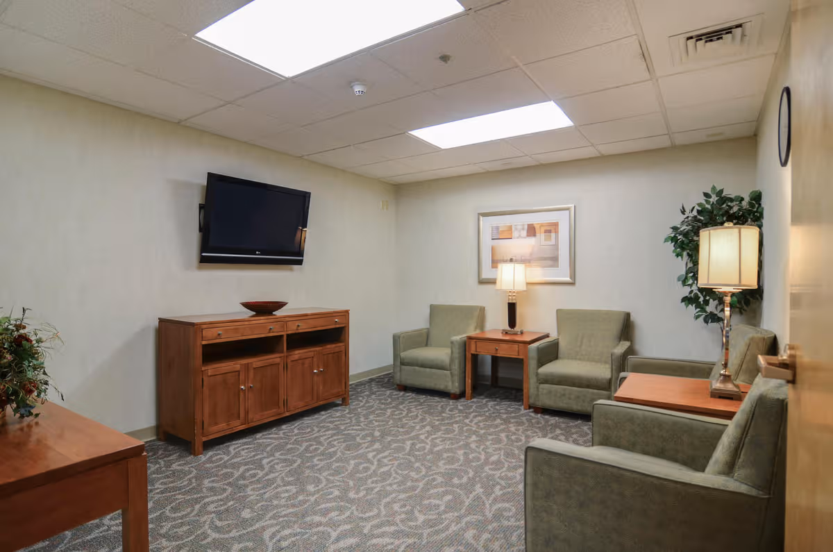 A quiet senior living lounge with green chairs, wooden tables, carpet, and a wall-mounted TV.