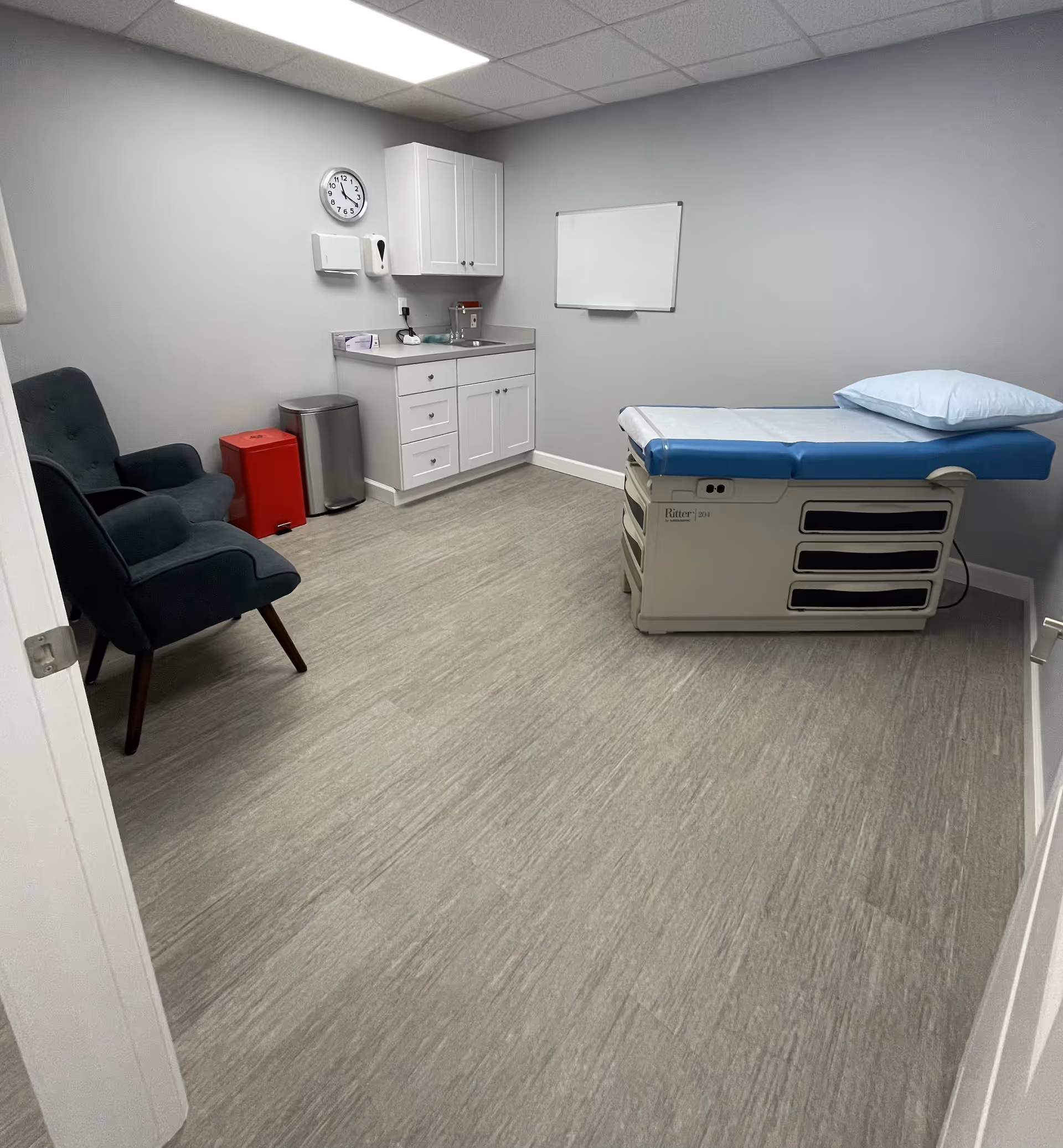 A medical examination room featuring an exam table, sink area, cabinets, a whiteboard, and two chairs.