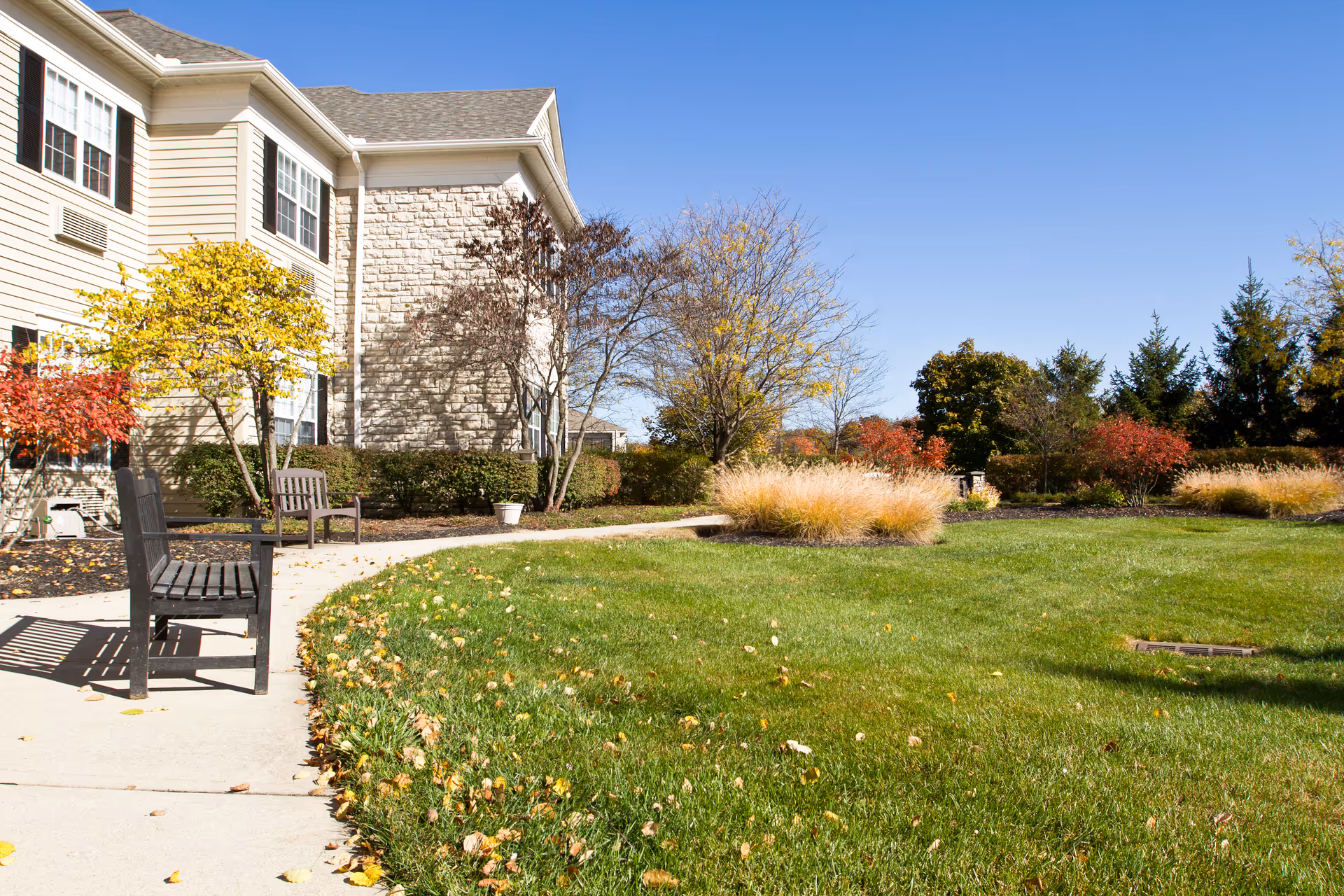Sunny exterior of Brookdale Muirfield with benches along a walkway and manicured lawn
