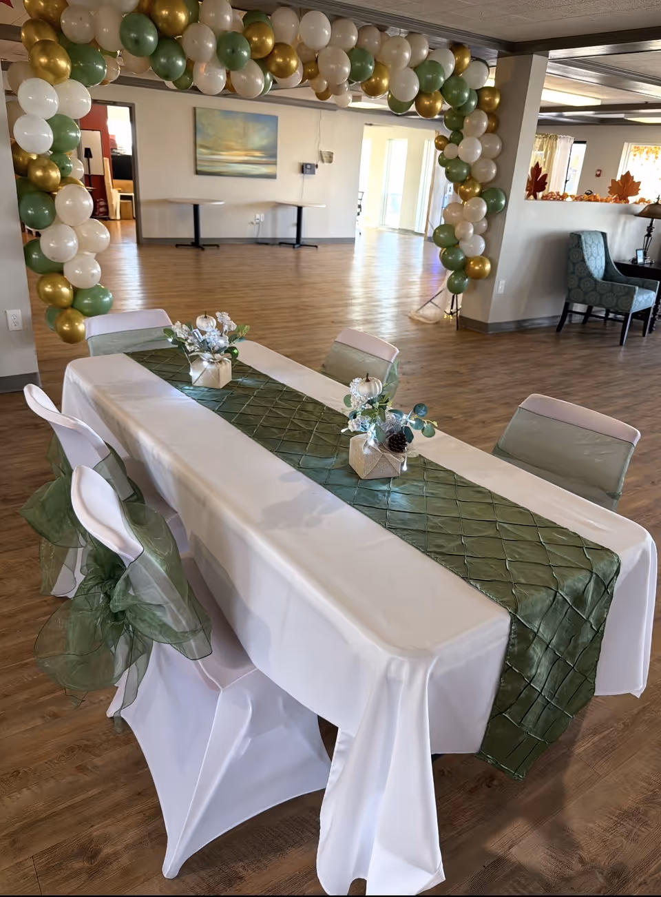 Indoor dining area with a long table, white linens, green runner, and balloon arch.
