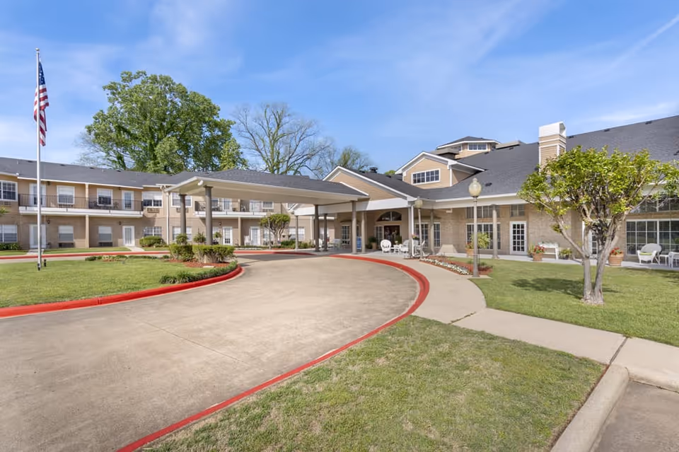 Exterior front entrance of a senior living facility with a covered porte-cochère, circular driveway, flagpole, and landscaped lawn.